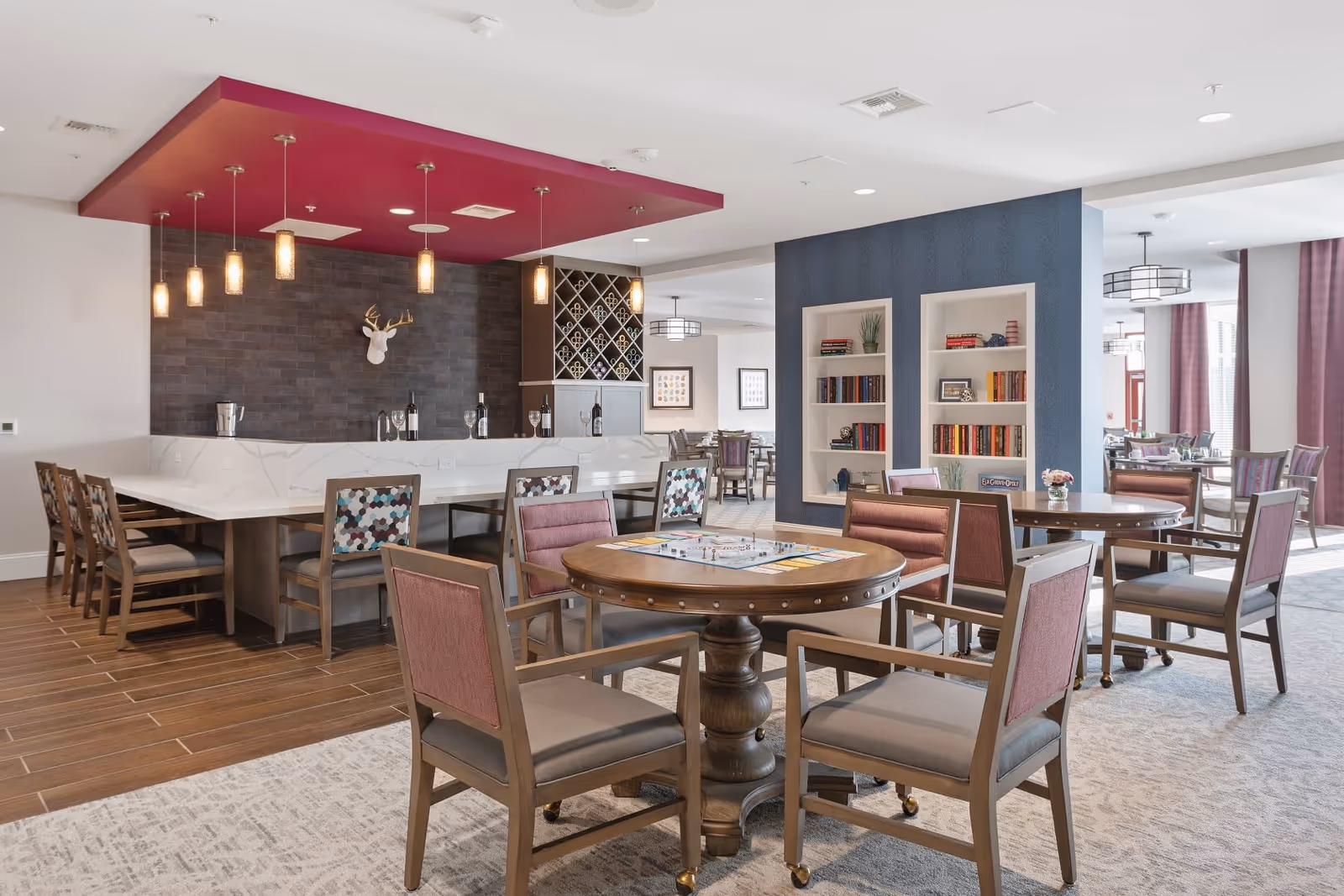 Communal dining and lounge area with round game tables, upholstered chairs, a marble-topped bar with wine rack, bookshelves, and pendant lighting.