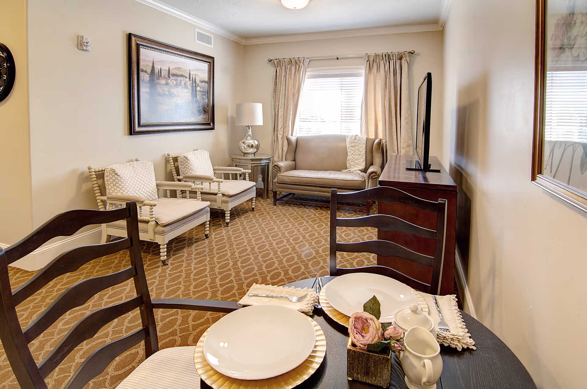 Living room area with a small dining table in the foreground, two armchairs and a loveseat by a window, a TV on a console, and framed artwork on the wall.
