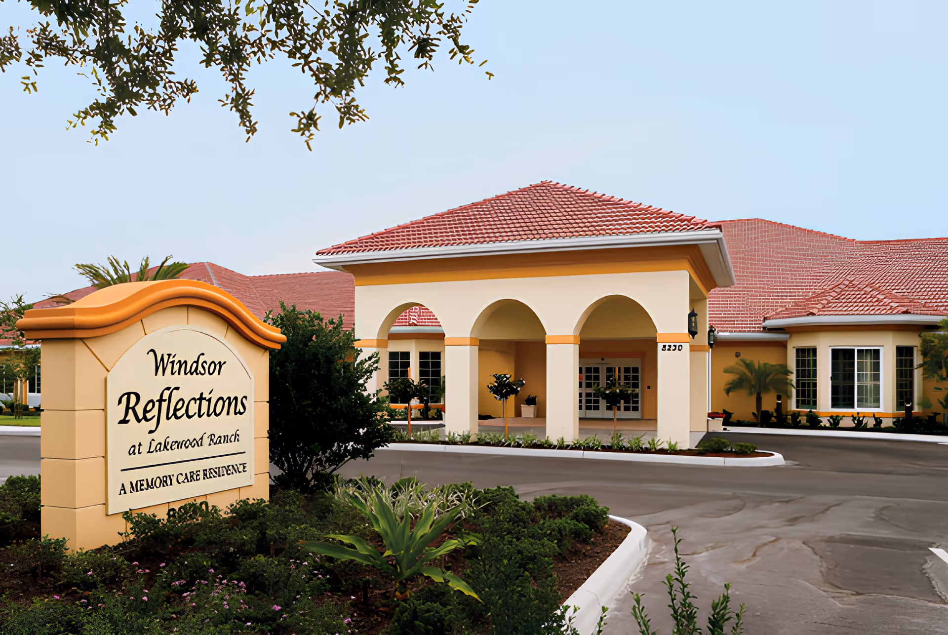 Exterior view of Windsor Reflections Memory Care residence at Lakewood Ranch, showing a building with a red tiled roof, cream and yellow walls, arched entrance, and landscaped greenery in front.