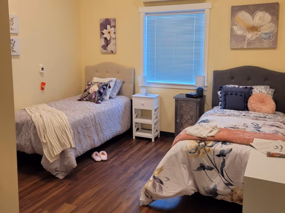 A cozy bedroom with two single beds, each with decorative pillows and floral bedding. Between the beds is a white nightstand with a lamp, and a window with closed blinds is centered on the wall behind. The room has wooden flooring and light-colored walls adorned with floral artwork. A pair of pink slippers is placed on the floor near one bed.