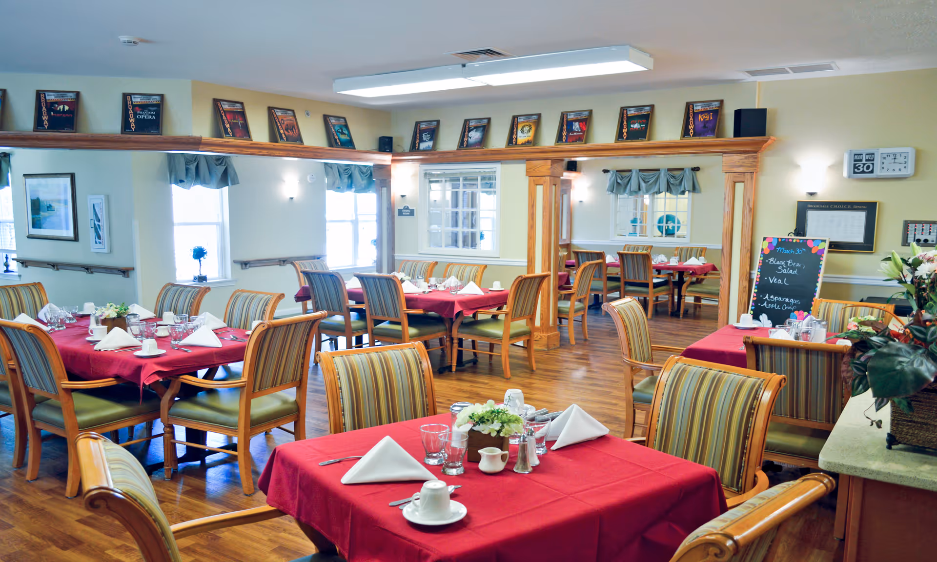 A bright dining room with several tables covered in red tablecloths, each set with white napkins, cups, glasses, and silverware. The room features wooden chairs with striped cushions, wooden flooring, and large windows with blue valances. There are framed pictures on the walls and a chalkboard menu displaying the day's meal. The space is well-lit with ceiling lights and wall sconces.