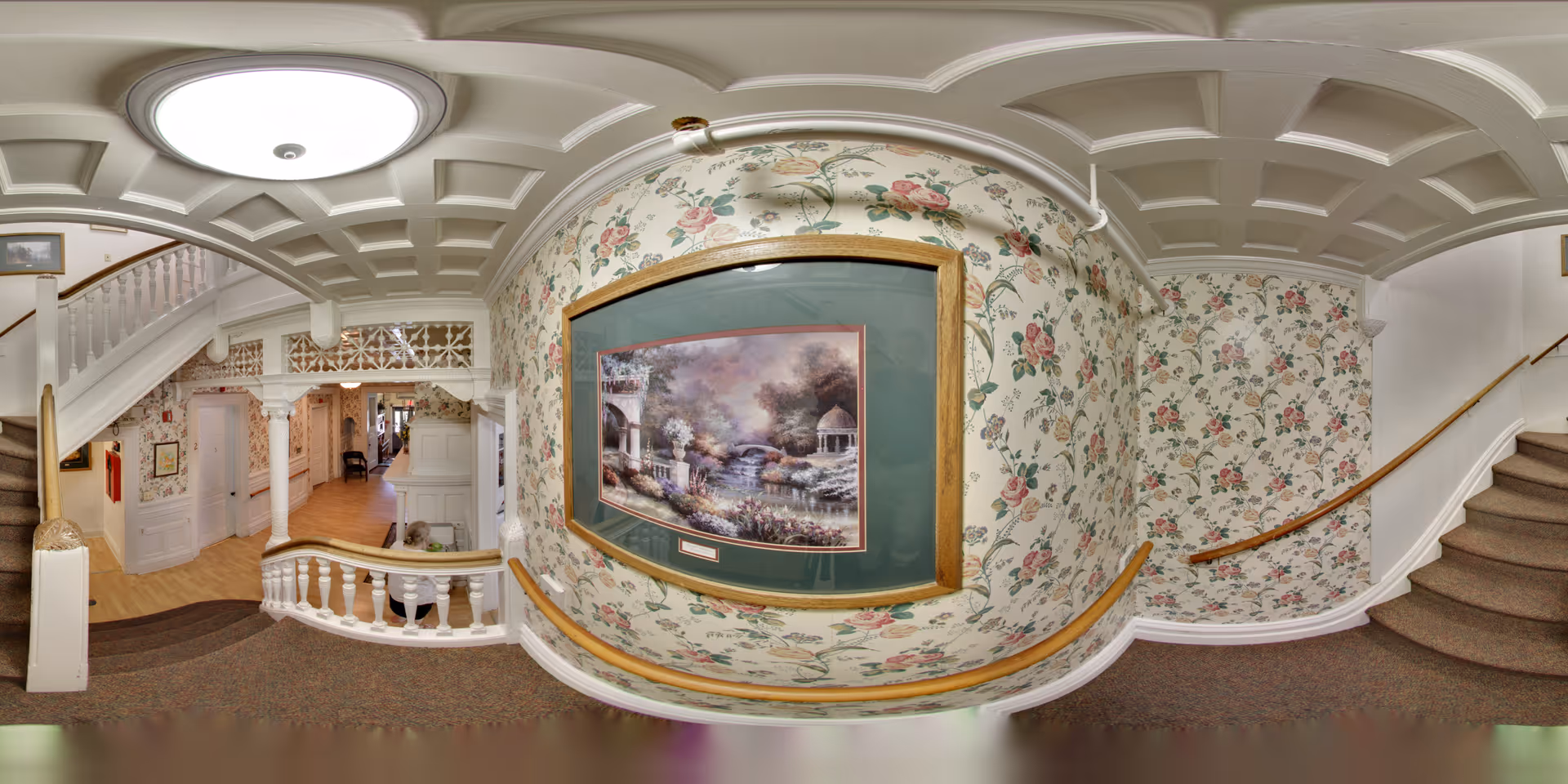Interior view of a senior living facility hallway with floral wallpaper, a framed painting of a garden scene, carpeted stairs with wooden handrails on both sides, and a white coffered ceiling with a round ceiling light. The hallway has wooden flooring and white decorative columns near the stairway.