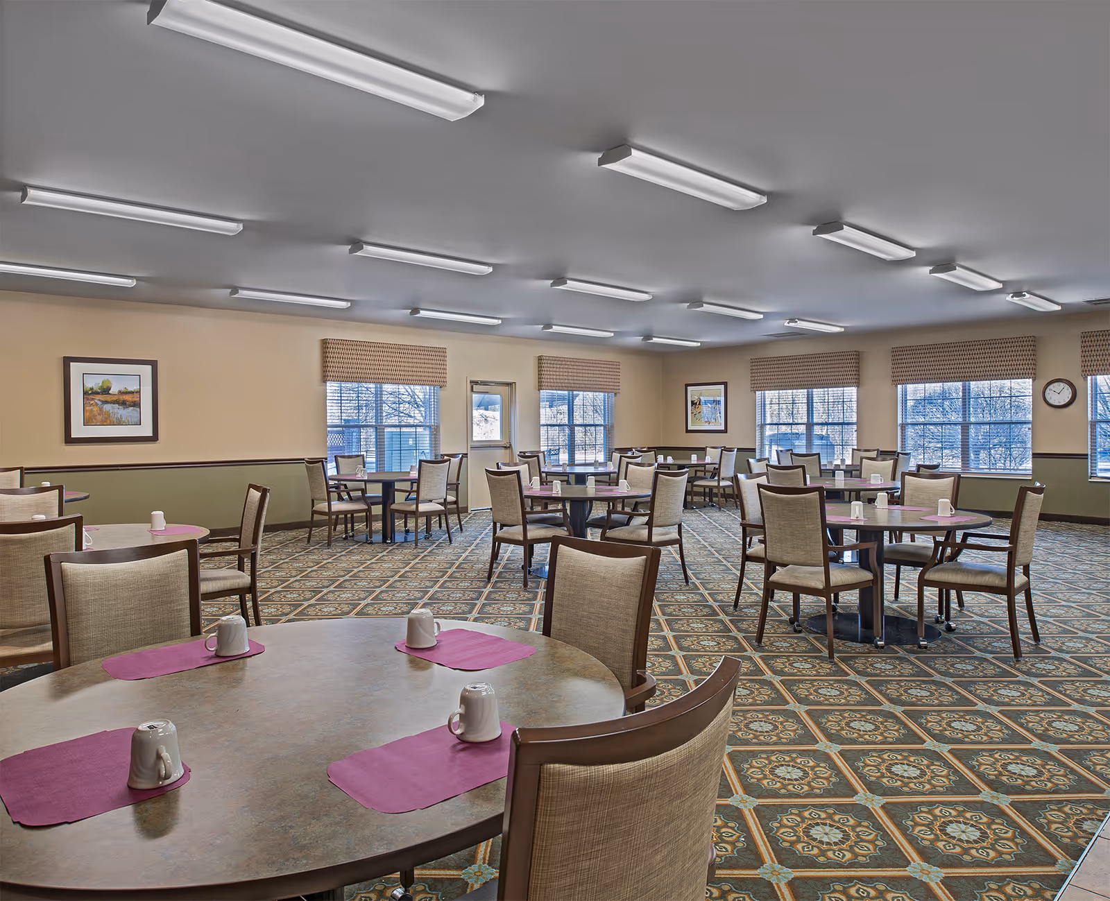 Large communal dining room with round tables and upholstered chairs on patterned carpet, each table set with placemats and mugs.