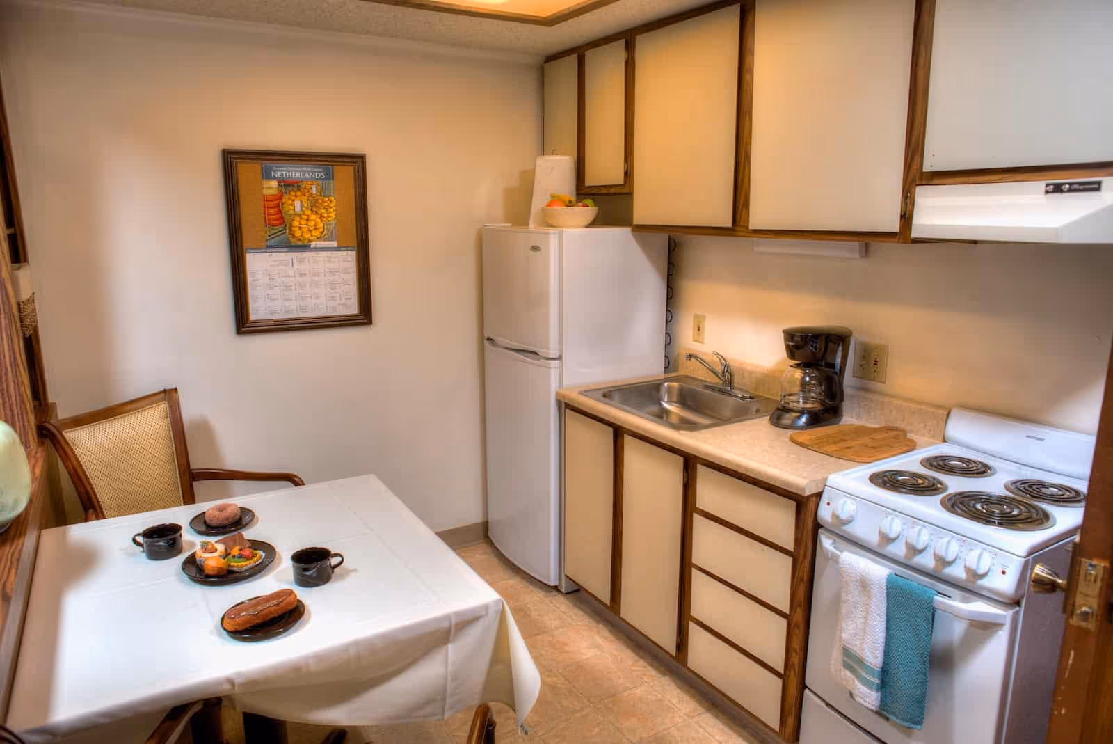 A small kitchen area in Springfield Assisted Living with a white refrigerator, a sink, a coffee maker, and a white electric stove with a towel hanging on the handle. There are wooden cabinets above and below the counter. A table covered with a white tablecloth is set with plates of pastries and two black coffee mugs. A chair with a woven backrest is positioned next to the table. A framed calendar hangs on the wall.