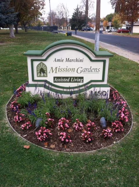 Entrance sign for Mission Gardens Assisted Living with a flower bed and greenery.