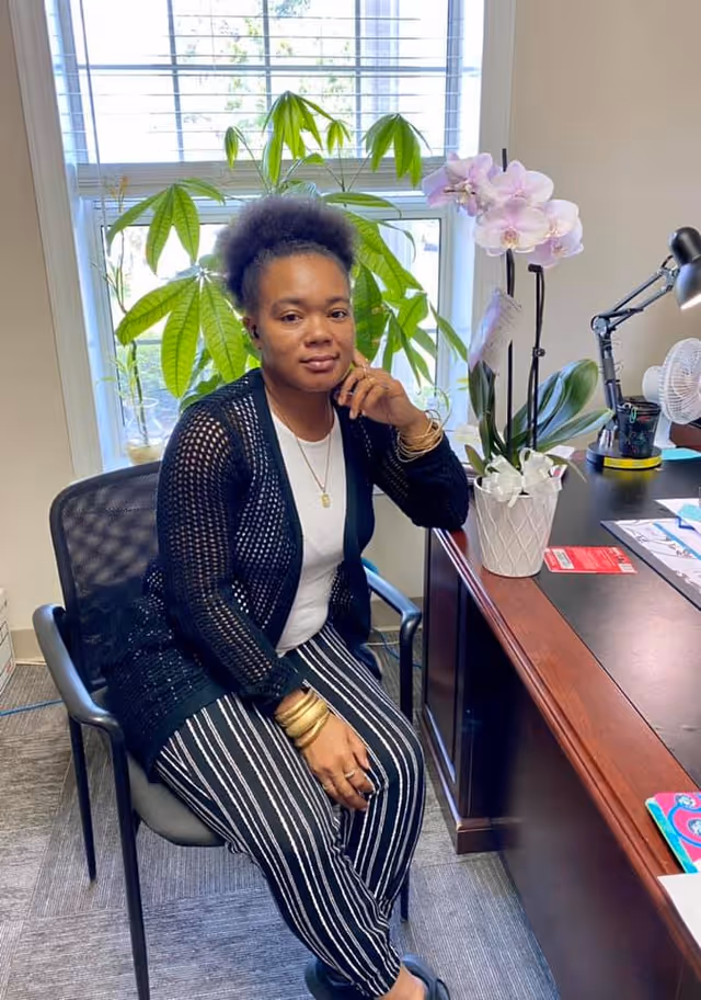 A woman with natural hair sits on a chair next to a wooden desk in an office. She is wearing a black open-knit cardigan, a white shirt, striped black and white pants, and gold bracelets. Behind her is a window with green plants visible outside. On the desk, there is a white orchid plant, a desk lamp, a small fan, and various papers and office supplies.