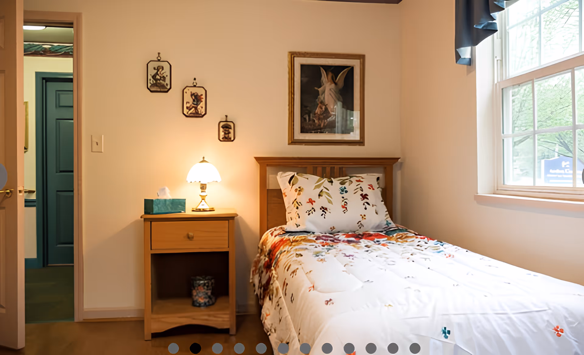A small, warmly lit single bedroom with a wooden bed, floral bedding, a nightstand with lamp and tissue box, and a window.