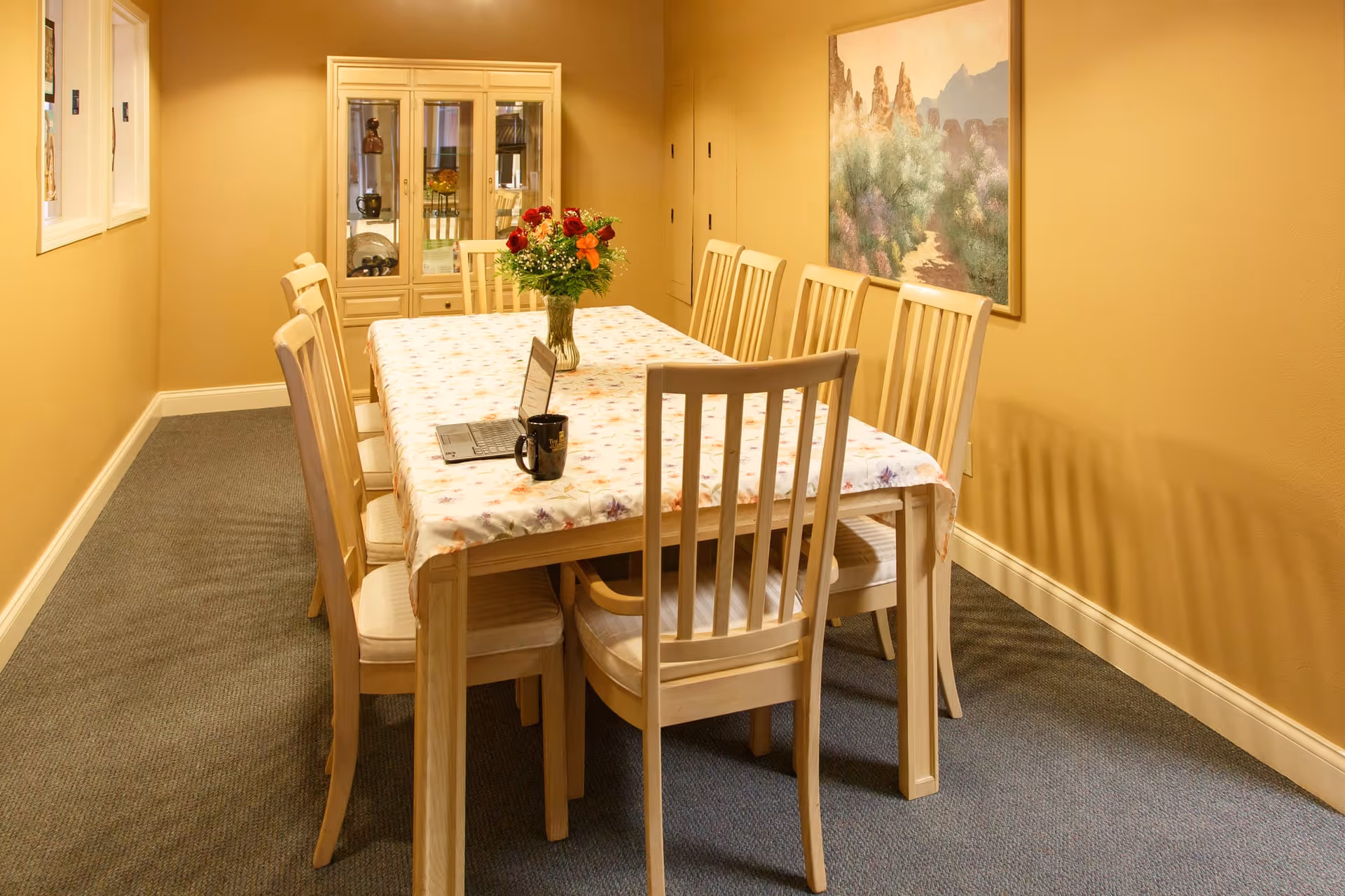 A dining room with a long rectangular wooden table covered with a floral tablecloth, surrounded by eight wooden chairs with cushioned seats. A vase with a bouquet of flowers is placed in the center of the table, along with a laptop and a black mug. The room has beige walls, a carpeted floor, a glass-front cabinet with decorative items, and a large landscape painting on the wall.