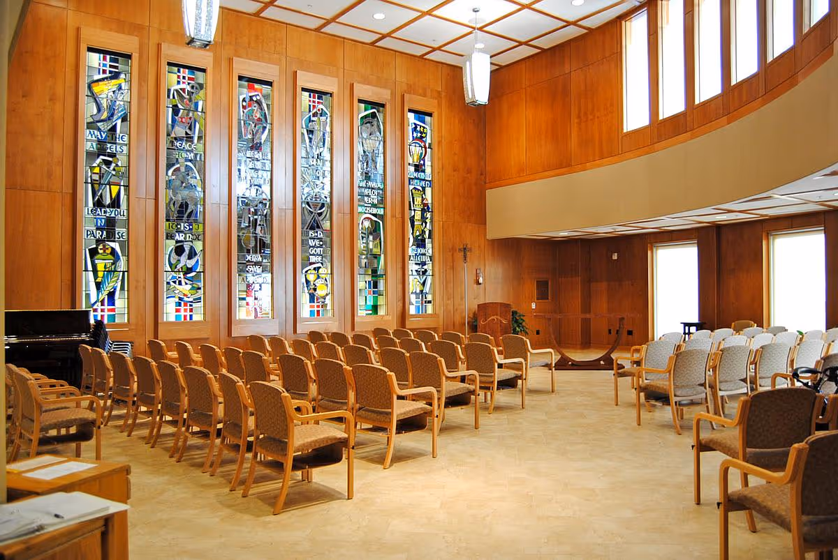 Interior view of a chapel or worship room with rows of wooden chairs arranged facing a podium. The walls are wooden paneled with tall stained glass windows depicting religious symbols. The ceiling has a grid pattern with hanging light fixtures, and there are large windows near the ceiling letting in natural light.