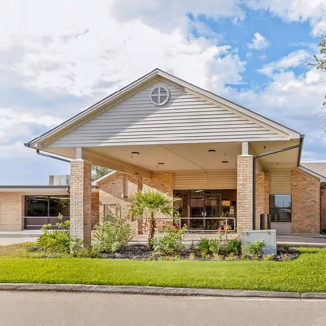 Front entrance of a single-story brick nursing facility with a covered porte-cochere, glass doors, and landscaped beds.
