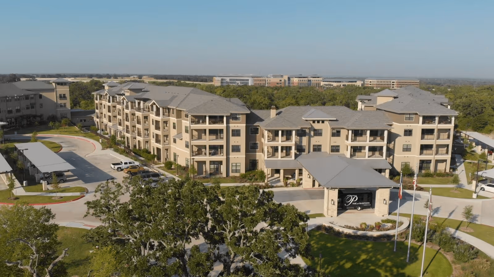 Aerial view of Parc Retirement Community at Traditions, TX, showing a large multi-story beige building with multiple balconies, a covered entrance with a sign, surrounding greenery, parking areas, and a clear sky.