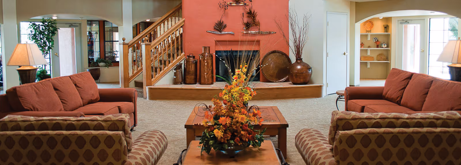 A cozy communal living room with matching sofas and armchairs arranged around a coffee table with a floral centerpiece, a fireplace, and a staircase in the background.