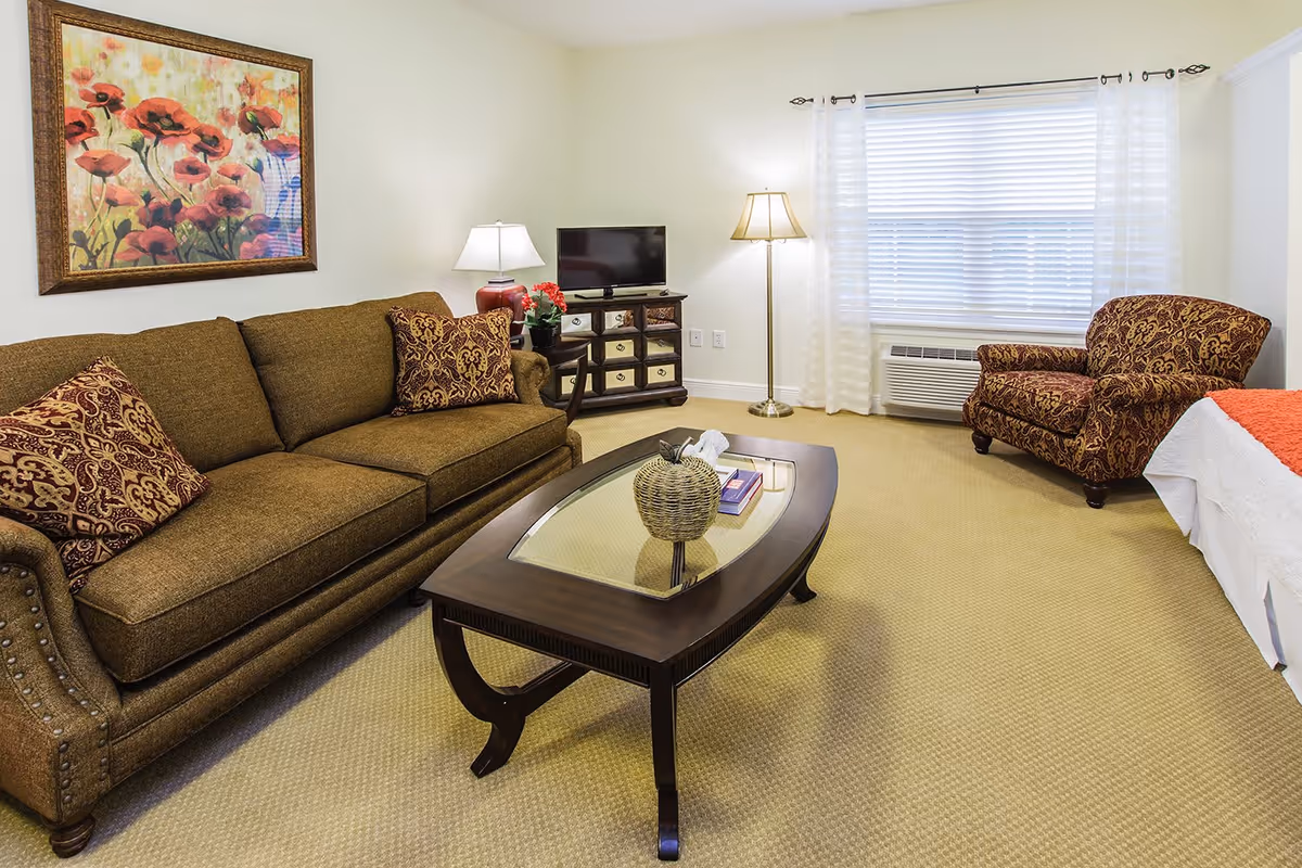 A cozy living room area in a senior living facility featuring a brown sofa with patterned cushions, a matching armchair, a wooden coffee table with a glass top and decorative items, a small TV on a wooden cabinet, two lamps, a large window with white blinds and sheer curtains, and a framed floral painting on the wall.