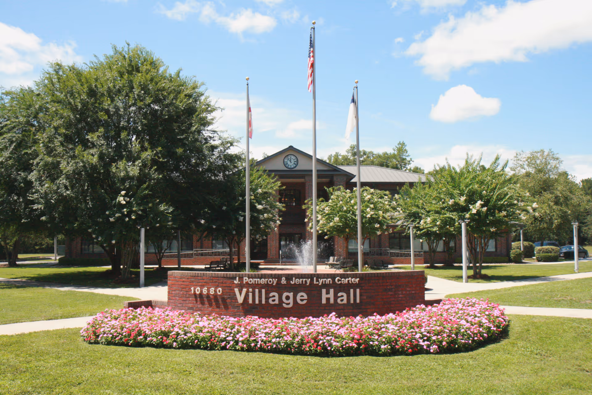 Front exterior view of J. Pomeroy & Jerry Lynn Carter Village Hall building with a brick sign surrounded by pink flowers, three flagpoles with flags, and trees on a sunny day.