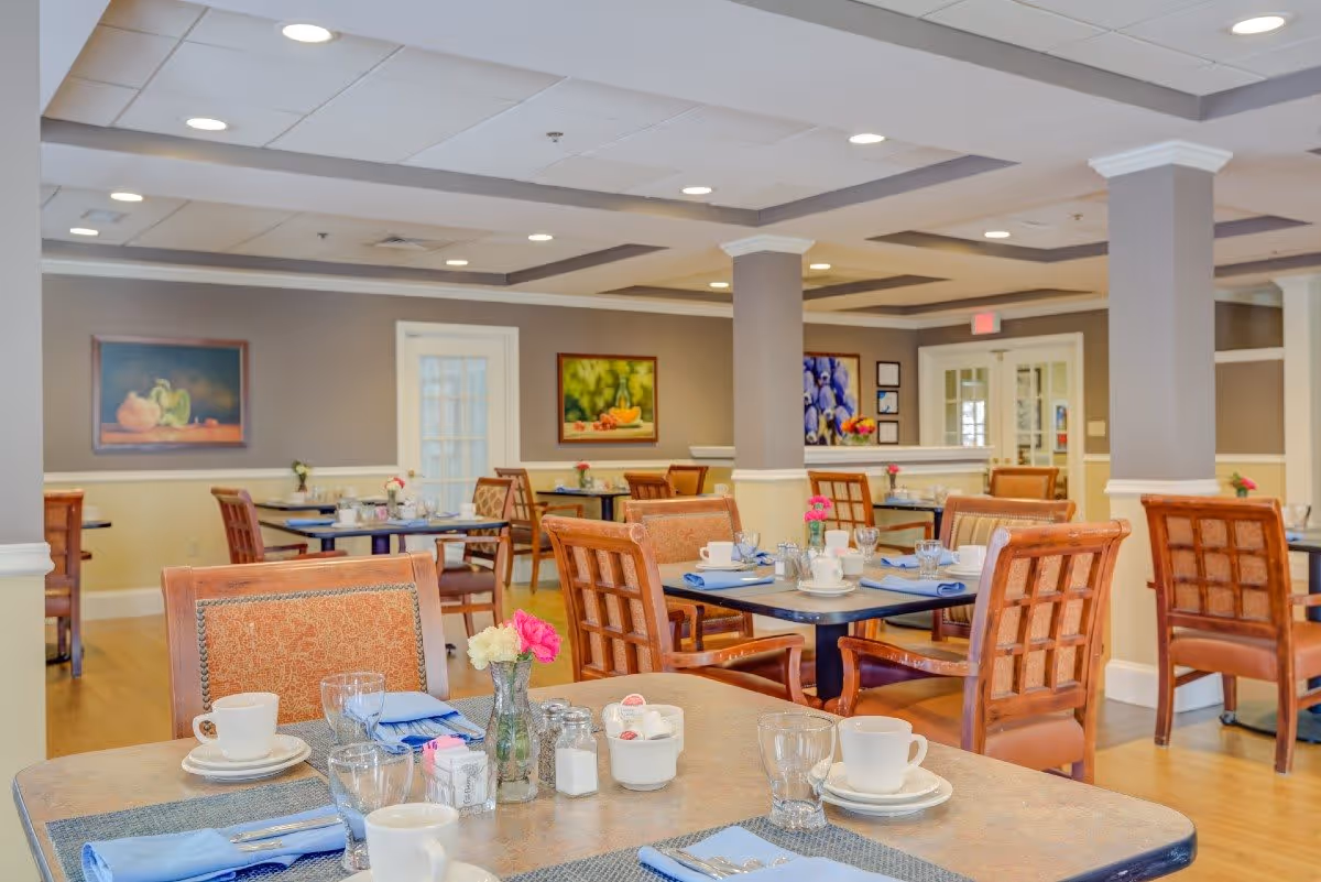 A bright and spacious dining room with multiple tables set for a meal. Each table has place settings including cups, glasses, napkins, and small flower vases. The room features wooden chairs with cushioned seats, soft lighting from recessed ceiling lights, and paintings on the walls. The overall decor is warm and inviting.