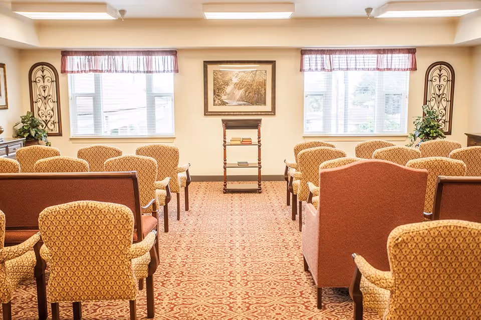 A well-lit room with rows of upholstered chairs arranged facing a small wooden stand with books. The room has two large windows with sheer curtains and valances, decorative wall art, and plants on either side near the windows. The carpet has a patterned design in warm tones.