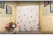 A bathroom with a shower curtain decorated with colorful butterflies. The walls are tiled halfway up in a light yellow color, and the upper walls are painted a light beige. There are framed pictures hanging on the walls and a basket with artificial flowers placed on a surface to the left.