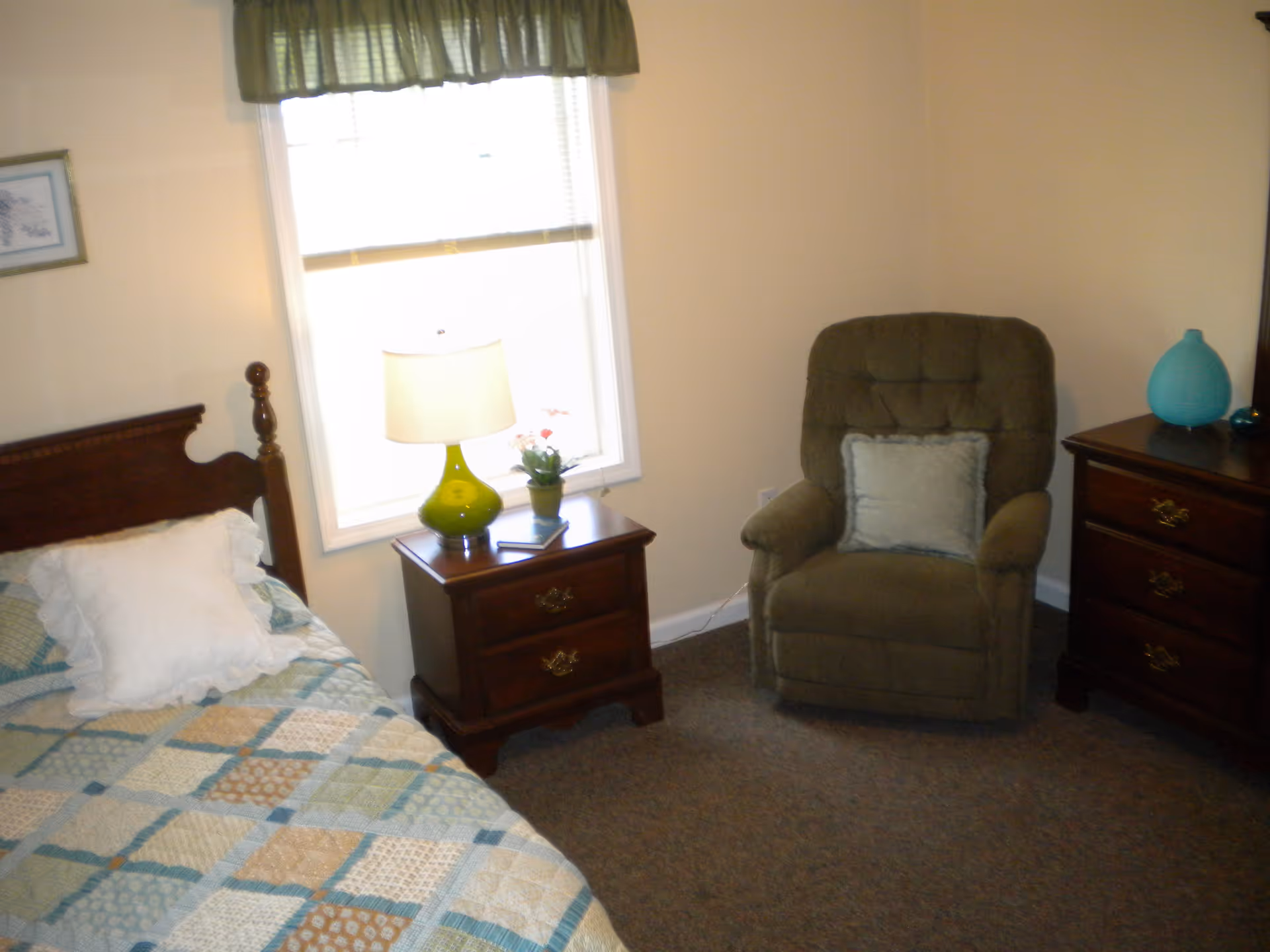 A bedroom with a single bed covered in a patchwork quilt and a white pillow. Next to the bed is a wooden nightstand with a green glass lamp and a small potted plant. There is a window with a green valance above the nightstand. Across from the bed is a cushioned armchair with a light blue pillow. To the right of the chair is a wooden dresser with a blue decorative vase on top.