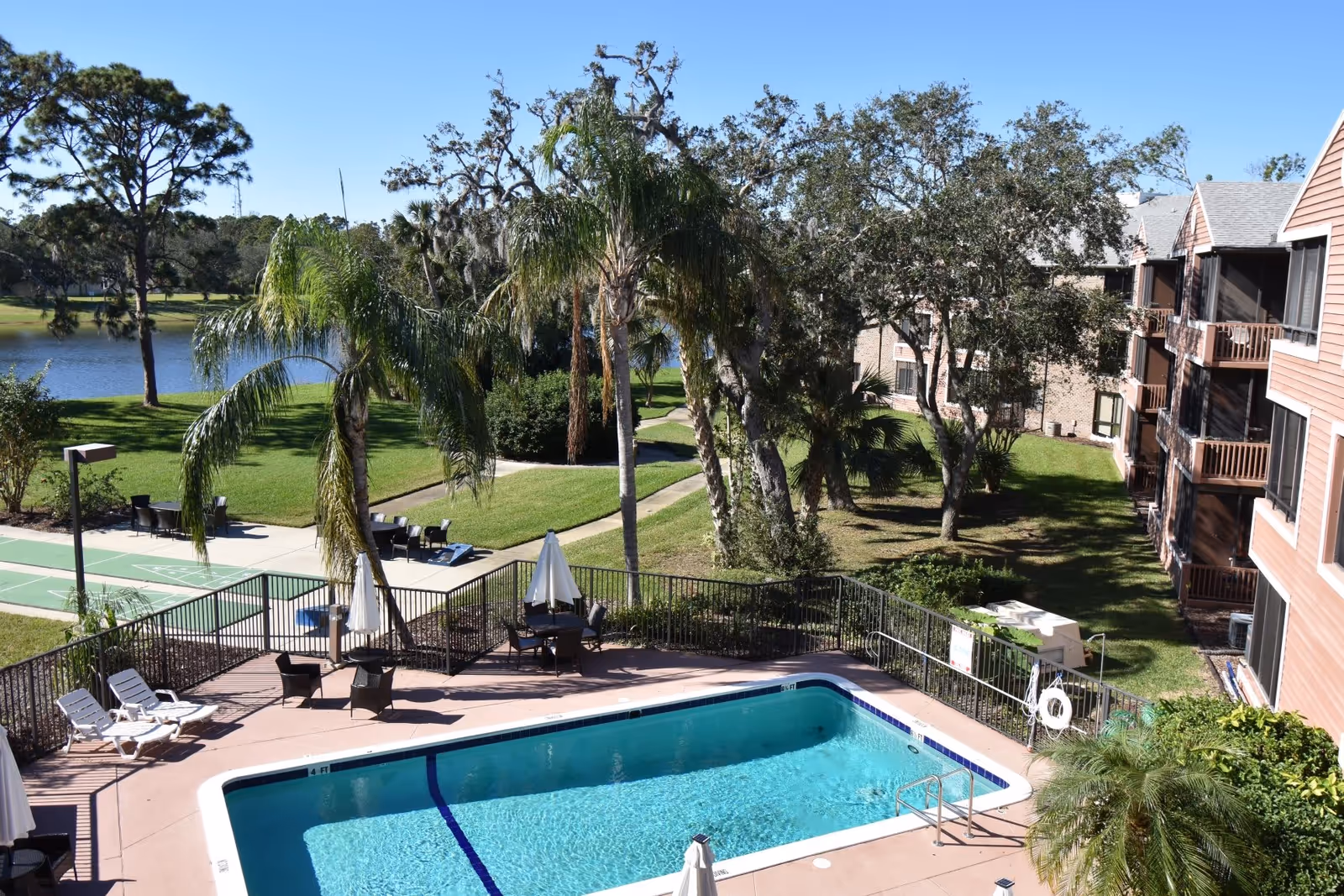 Outdoor swimming pool area with lounge chairs, tables with umbrellas, and a shuffleboard court next to a lake. Surrounding the pool are palm trees and other greenery, with a multi-story residential building on the right side.
