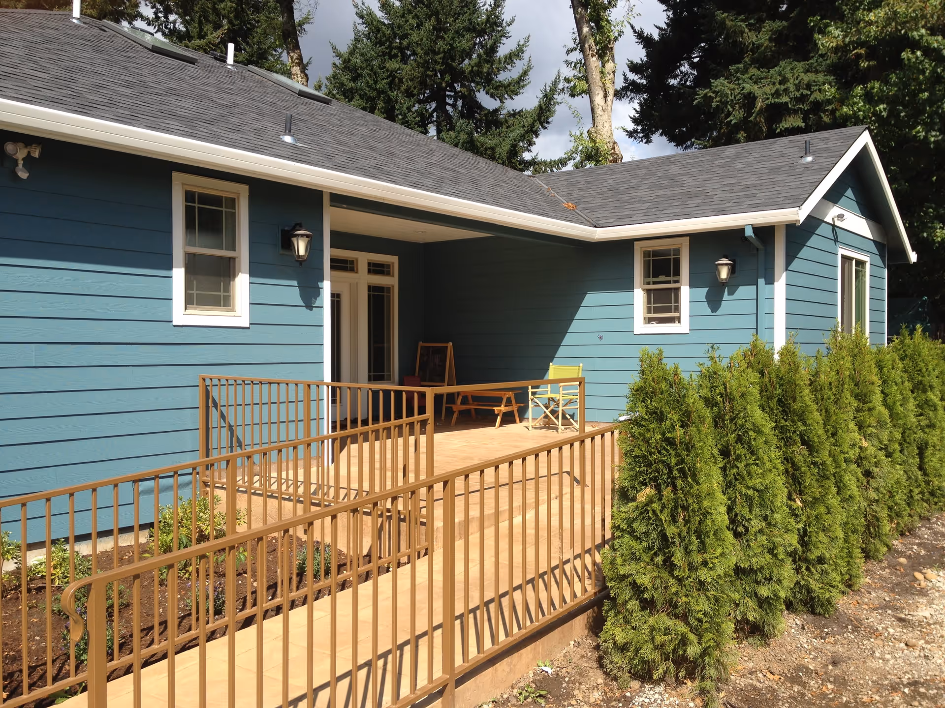 Exterior view of a blue single-story building with white trim, featuring a wooden ramp with metal railings leading to a covered porch area. There are two windows and a door on the building, outdoor wall-mounted lights, and a row of green shrubs along the side of the ramp. Trees and a partly cloudy sky are visible in the background.
