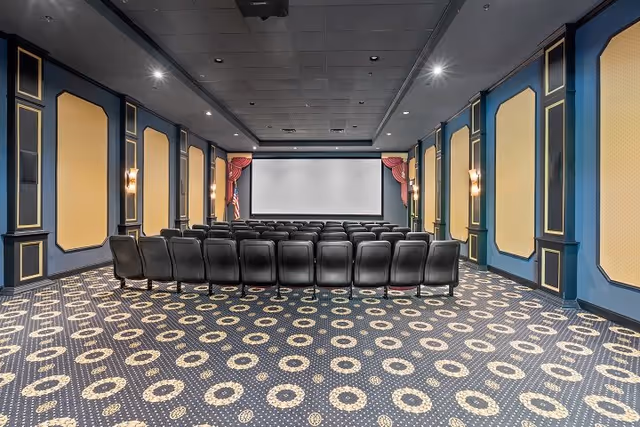Interior view of a theater-style room with rows of black chairs facing a large white projection screen. The room has blue walls with gold-trimmed panels and wall sconces, an American flag to the left of the screen, and a patterned carpet with circular designs.