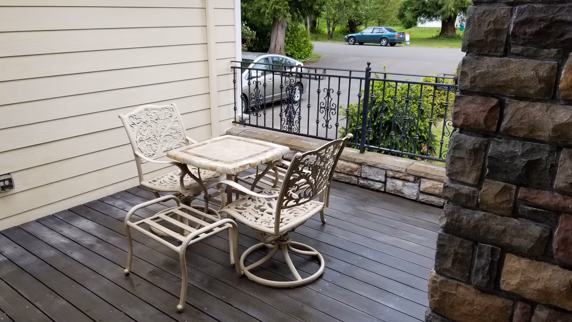 Outdoor patio area with a small square table and three ornate metal chairs on a wooden deck. The patio is adjacent to a beige siding wall and a stone pillar, with a black wrought iron railing overlooking a parking area with two parked cars and greenery in the background.