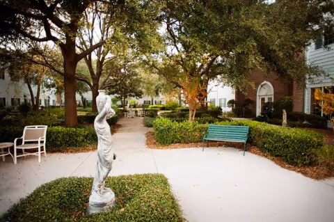 Outdoor courtyard area with a statue of a woman holding a jug, surrounded by trees, bushes, benches, and white buildings in the background.