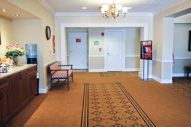 Carpeted entry hallway of a senior living community with a bench, water cooler, service counter, and double doors.
