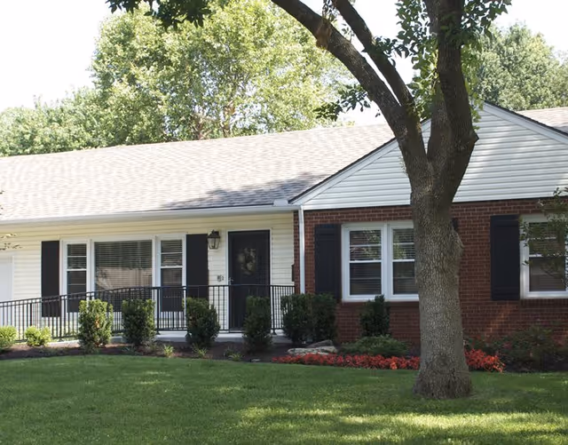 Exterior view of a single-story senior living facility building with white siding and red brick, black shutters on windows, a black front door, a metal railing along the entrance ramp, surrounded by green grass, bushes, a tree, and flower beds.