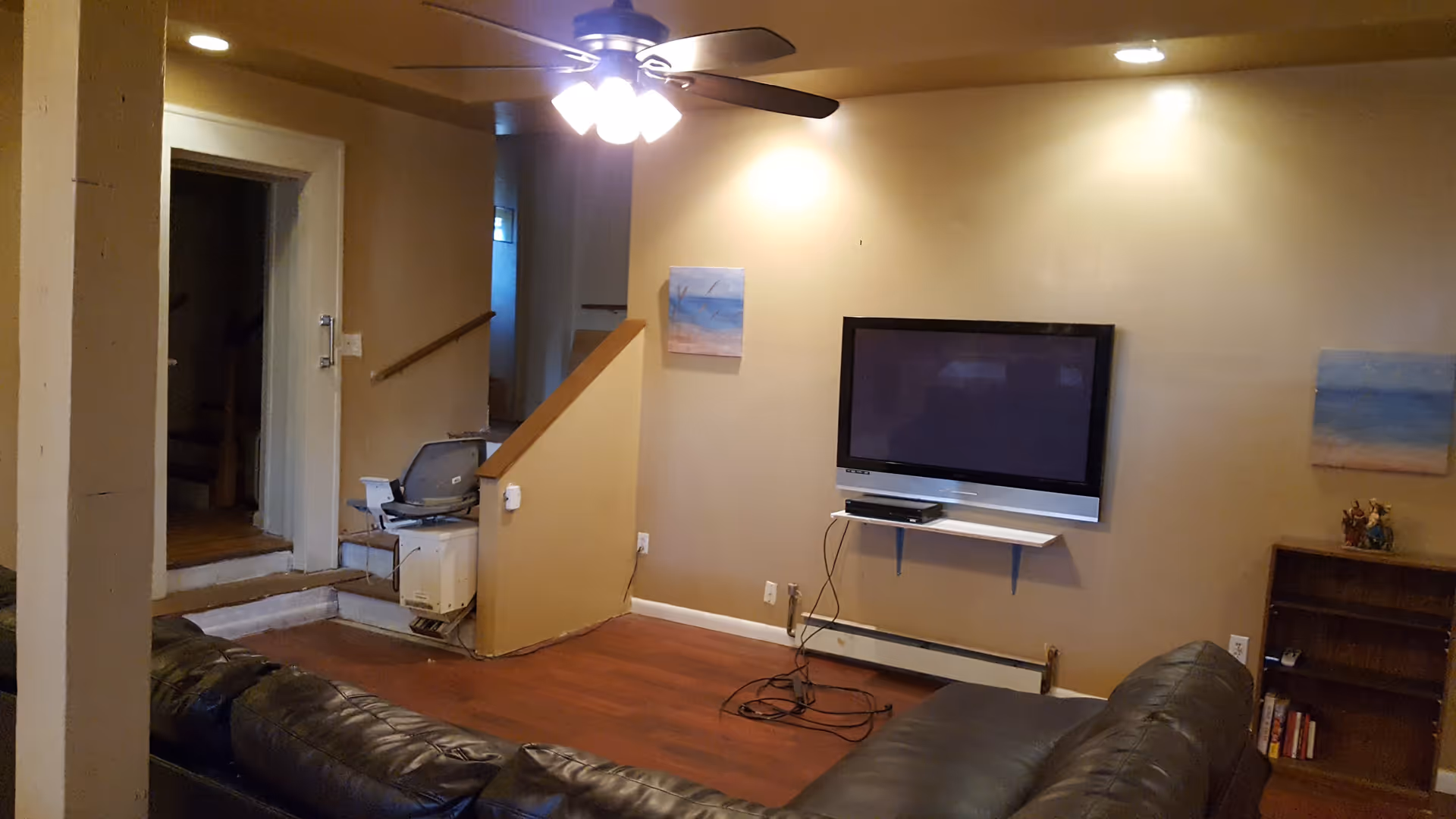 Interior living room with a black leather sectional sofa, a wall-mounted flat screen TV with cables hanging below, a small wooden bookshelf with some books and figurines, two small paintings on the wall, a ceiling fan with lights, and a stairlift installed on a short staircase leading to another room.