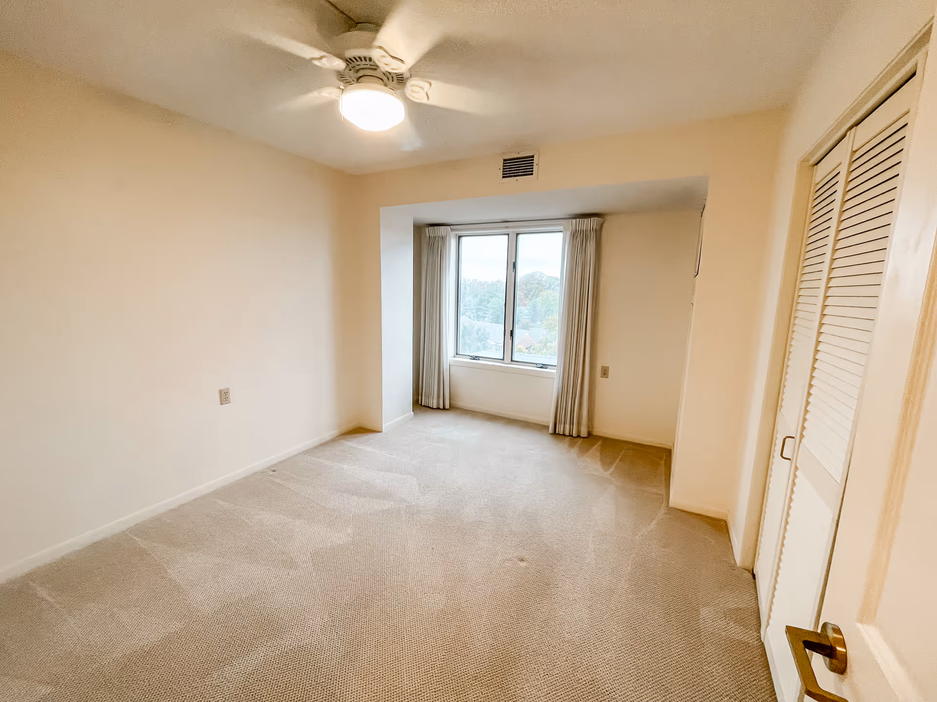 Empty bedroom with beige carpet, cream-colored walls, a ceiling fan with light, a window with curtains overlooking trees outside, and a closed closet with louvered doors.