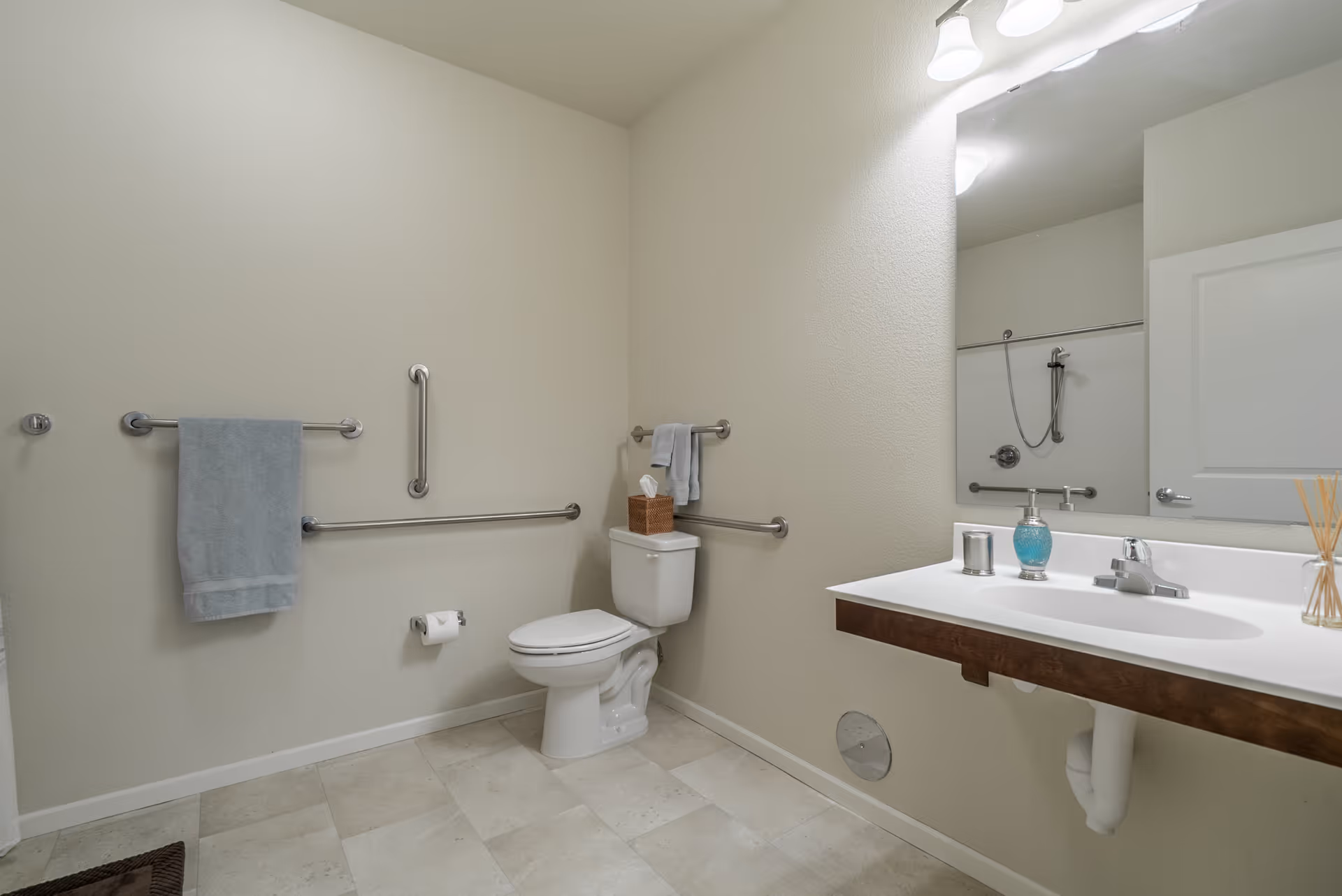 A clean and accessible bathroom with beige walls and tiled floor. The room features a white toilet with grab bars on the walls around it, a towel rack with a blue towel, a large mirror above a white sink with a wooden base, and a shower area visible in the mirror reflection. The sink countertop holds a soap dispenser, a small container, and reed diffusers. The bathroom is well-lit with wall-mounted lights above the mirror.