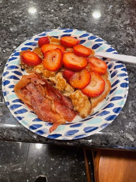Paper plate with a waffle topped with sliced strawberries and a side of bacon on a dark granite countertop.