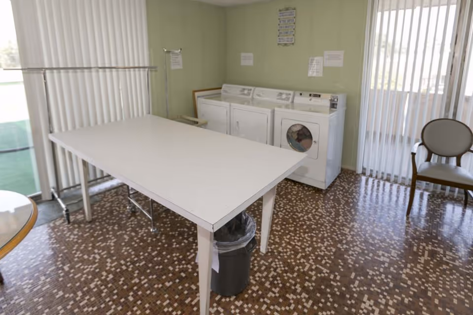 Laundry room with a white folding table in the center, a trash can underneath, three white laundry machines against a light green wall, a chair to the right, and vertical blinds covering windows on two sides.