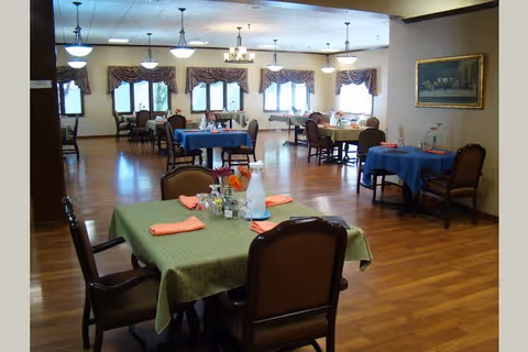 A spacious dining room with multiple tables covered in green, blue, and yellow tablecloths. Each table is set with napkins, glasses, and small flower arrangements. The room has wooden flooring, large windows with valances, and ceiling lights providing warm illumination. There is a framed painting on the right wall.