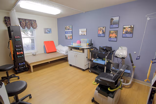 A therapy or rehabilitation room with exercise equipment including a recumbent bike, weights on a rack, and a padded therapy table. The room has light wood flooring, a window with floral curtains, and several framed photos on a blue accent wall.