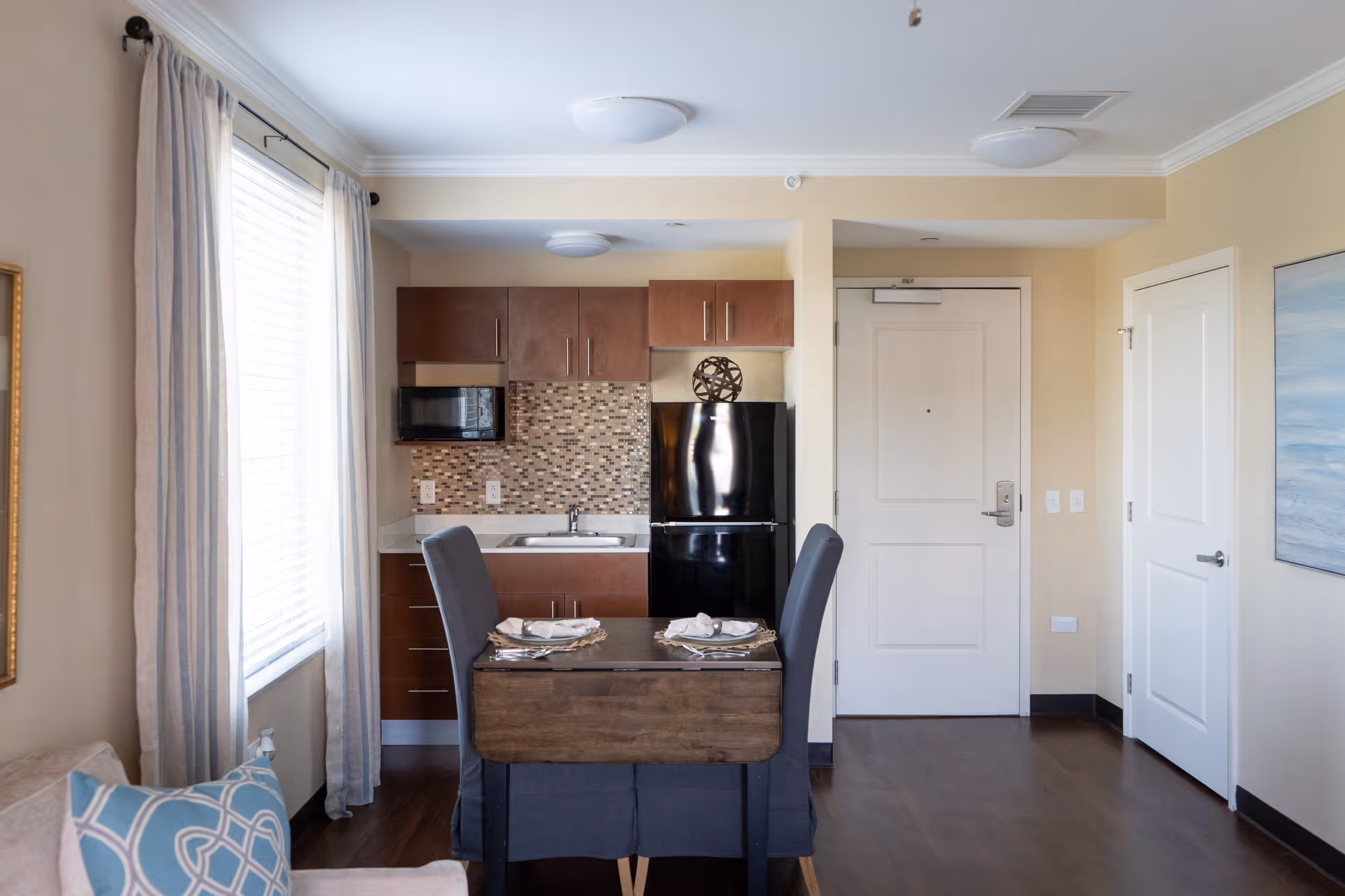 A small kitchen and dining area in a senior living facility apartment. The kitchen has brown cabinets, a black refrigerator, a microwave, and a mosaic tile backsplash. In front of the kitchen is a wooden dining table set for two with gray upholstered chairs. To the left is a window with light curtains, and to the right are two white doors and a painting on the wall.