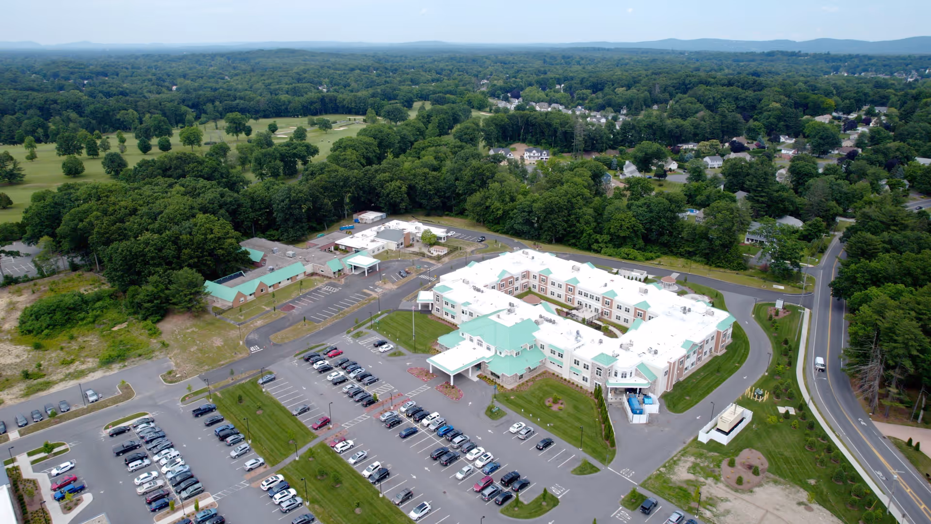 Aerial view of East Longmeadow Memory Care Assisted Living facility showing a large building with white roofs and green accents, surrounded by parking lots filled with cars, green lawns, trees, and nearby residential areas under a clear sky.