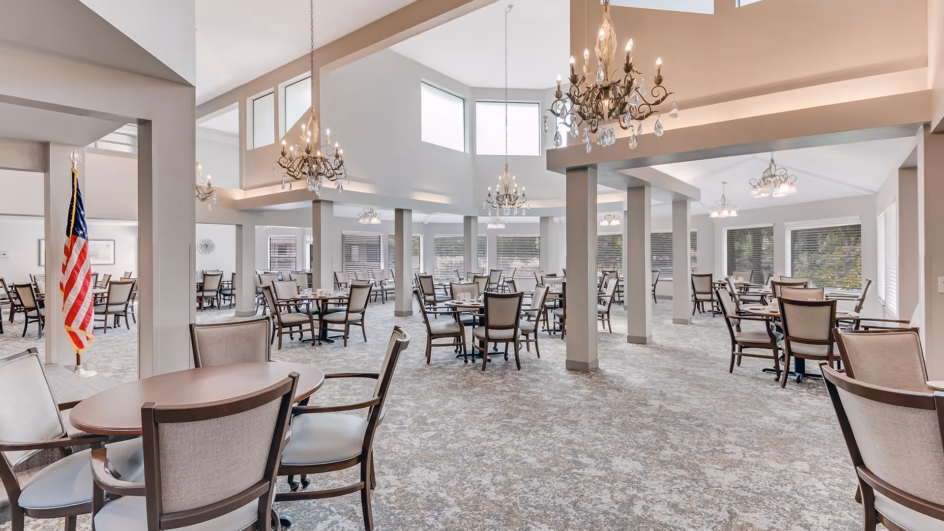 A spacious dining room with multiple round tables and cushioned chairs arranged neatly. The room features large windows allowing natural light to fill the space, elegant chandeliers hanging from the ceiling, and an American flag displayed near one of the pillars. The carpet has a subtle patterned design, and the walls are painted in light neutral tones.