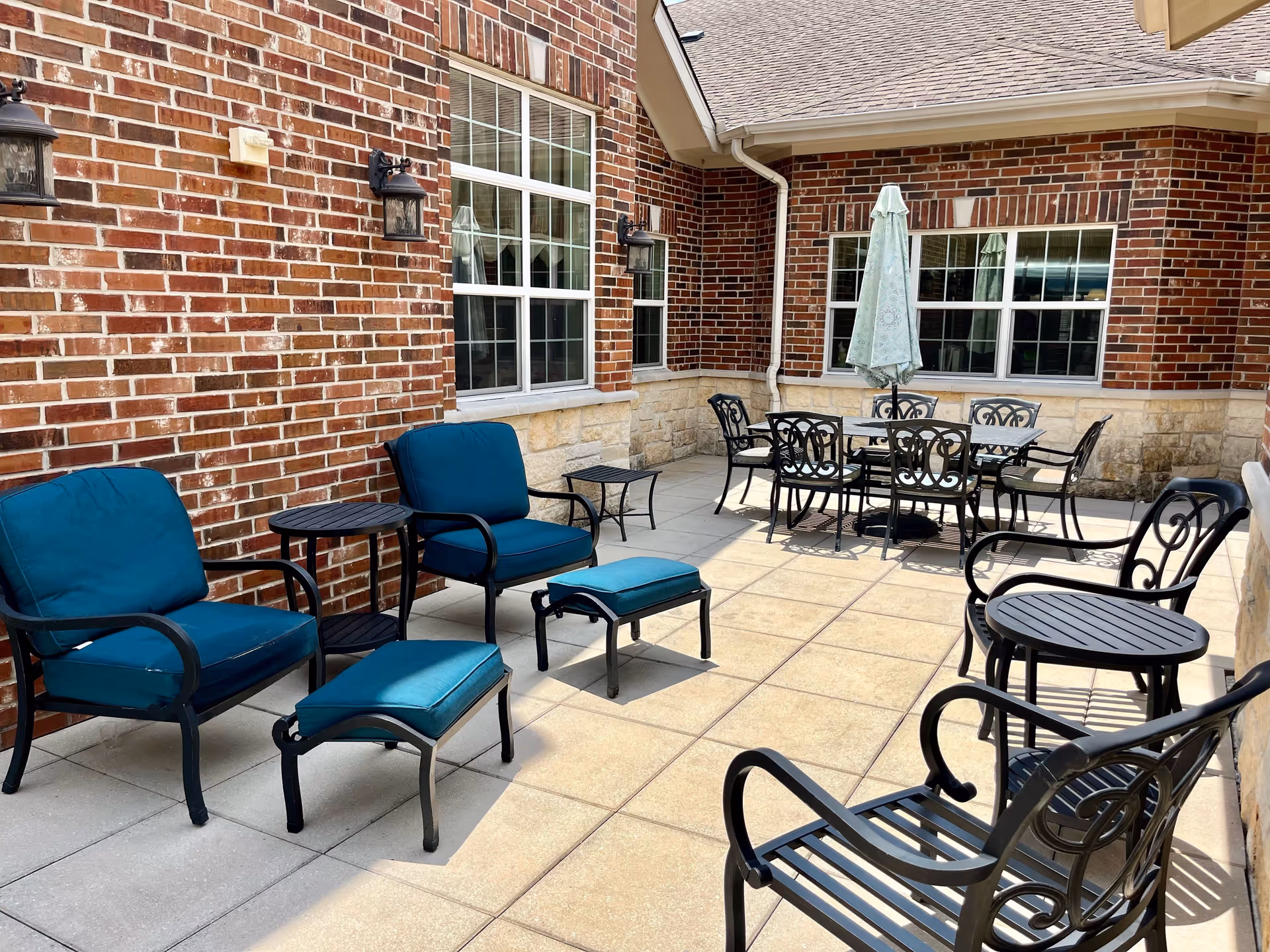 Outdoor patio area with brick walls and large windows. The patio is furnished with black metal chairs, tables, and blue cushioned armchairs with matching footrests. There is a closed umbrella over a dining table set in the background.