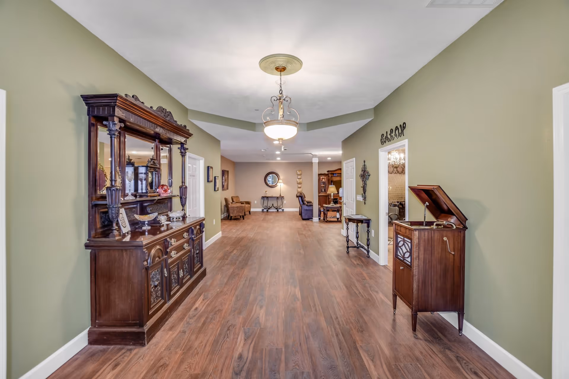 A long hallway with wooden flooring and green walls featuring antique wooden furniture including a large cabinet on the left and a vintage record player on the right. The hallway leads to a sitting area with chairs and a small table against a beige wall. A chandelier hangs from the ceiling, and there is a doorway labeled 'SALOON' on the right side.