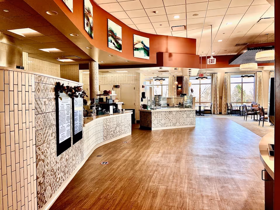 Interior view of a dining area in Monarch Landing featuring a curved counter with decorative tile patterns, menu boards, and a serving station. The space has wooden flooring, large windows with curtains allowing natural light, and several tables and chairs arranged for dining.