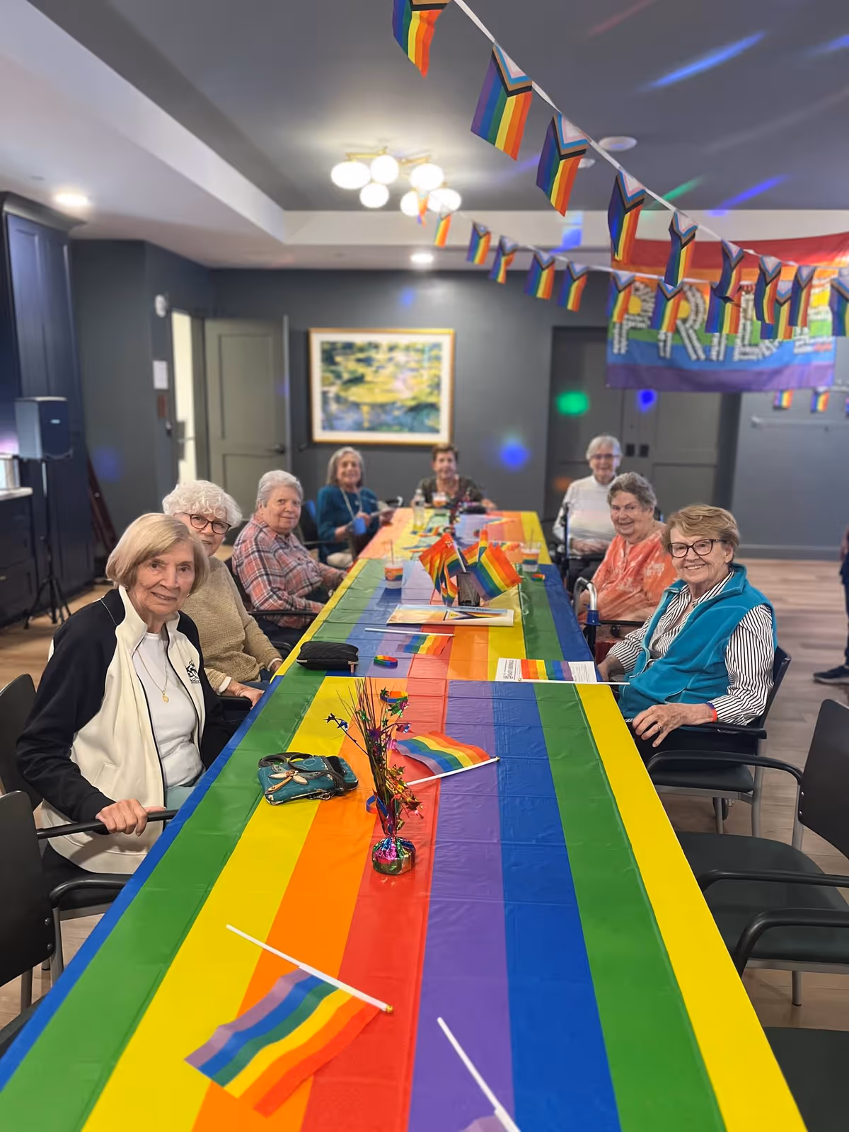 A group of older adults seated around a long table covered with rainbow Pride decorations in a communal dining/activity room.