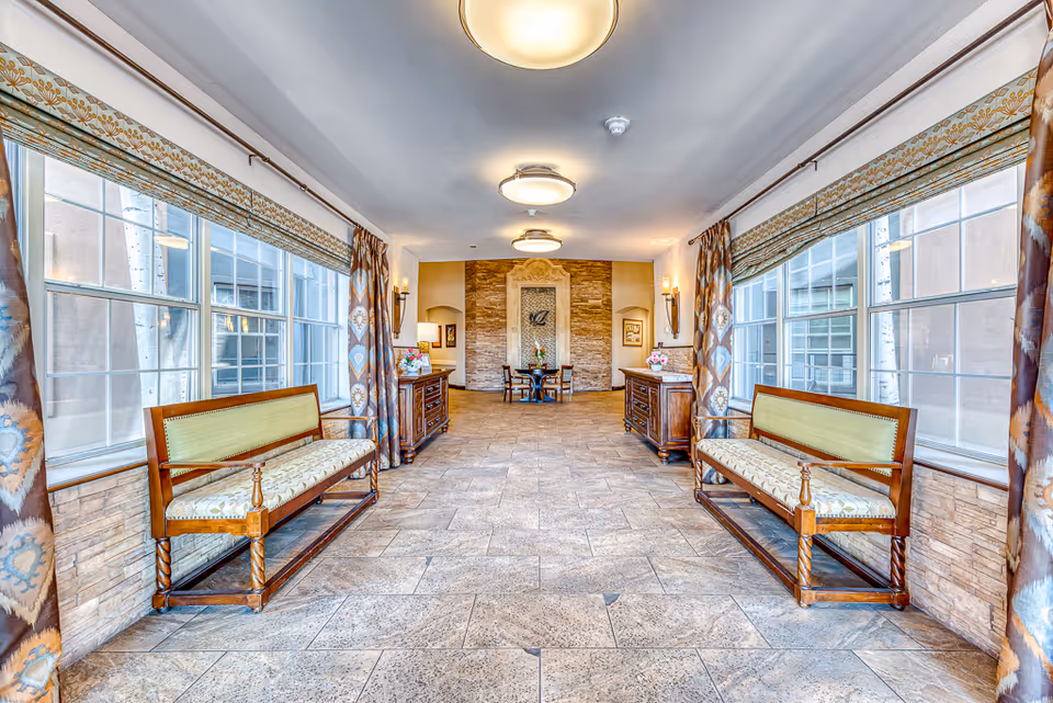 A bright and spacious interior hallway with large windows on both sides covered with patterned curtains. There are two wooden benches with cushioned seats facing each other along the windows. At the end of the hallway, there is a small round table with chairs and a decorative stone wall with an artistic feature. The floor is tiled with a stone pattern, and the ceiling has round light fixtures.
