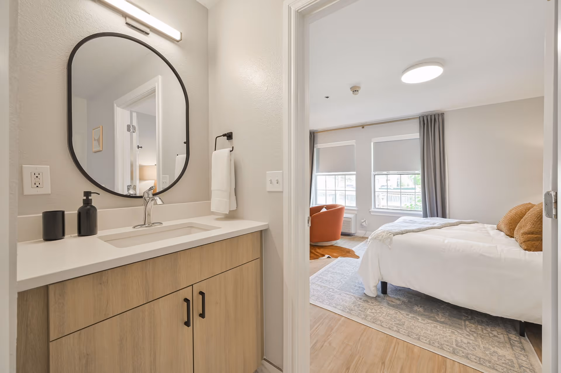 View of a modern bathroom vanity with a large round mirror, a sink, and soap dispensers on the countertop. Through an open door, a bedroom is visible with a bed, pillows, a patterned rug, two windows with blinds, and an orange armchair near the windows.