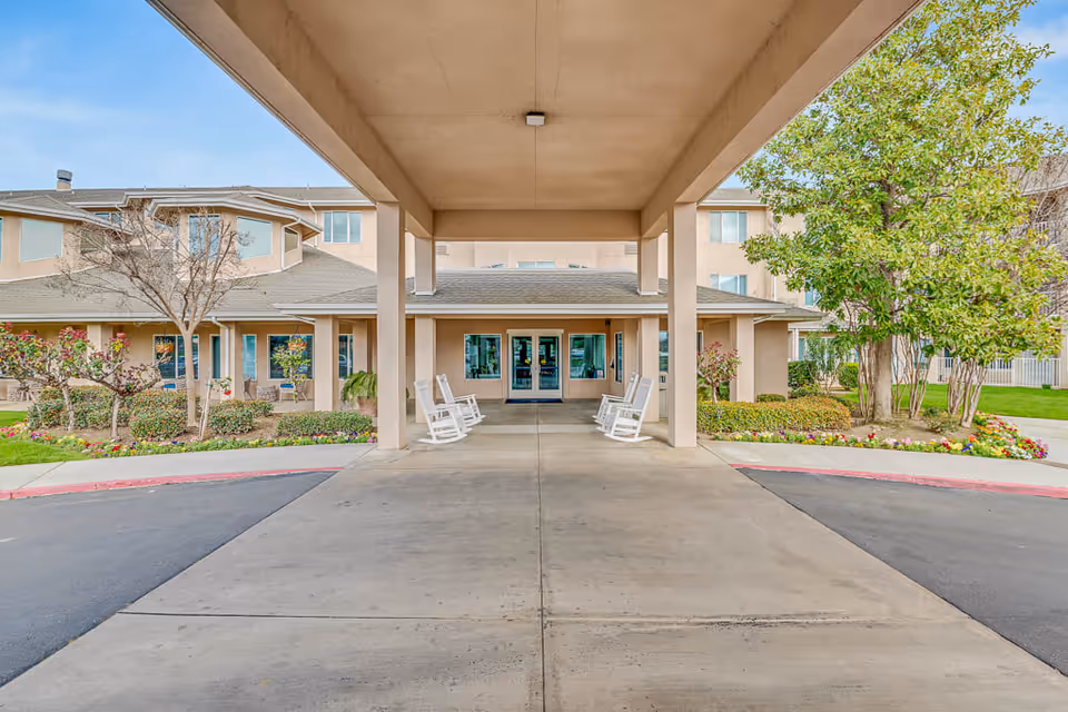 Covered entrance driveway of a senior living facility with white rocking chairs on either side, surrounded by landscaped bushes, flowers, and trees, leading to glass double doors of a beige building under a clear sky.