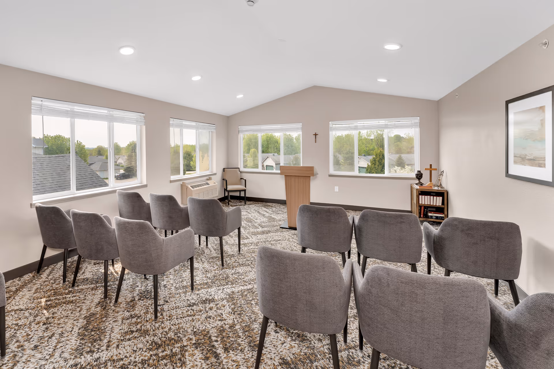 Bright meeting/chapel room with rows of gray chairs facing a wooden lectern and windows.