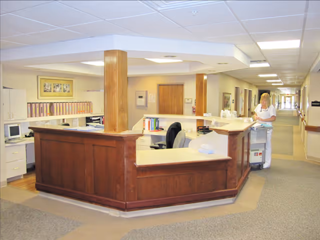 Reception desk area in a senior living facility with wooden paneling and a white countertop. A staff member in white uniform is standing near a cart in a long hallway with doors on both sides. The area is well-lit with ceiling lights and has beige walls and carpeted floors.