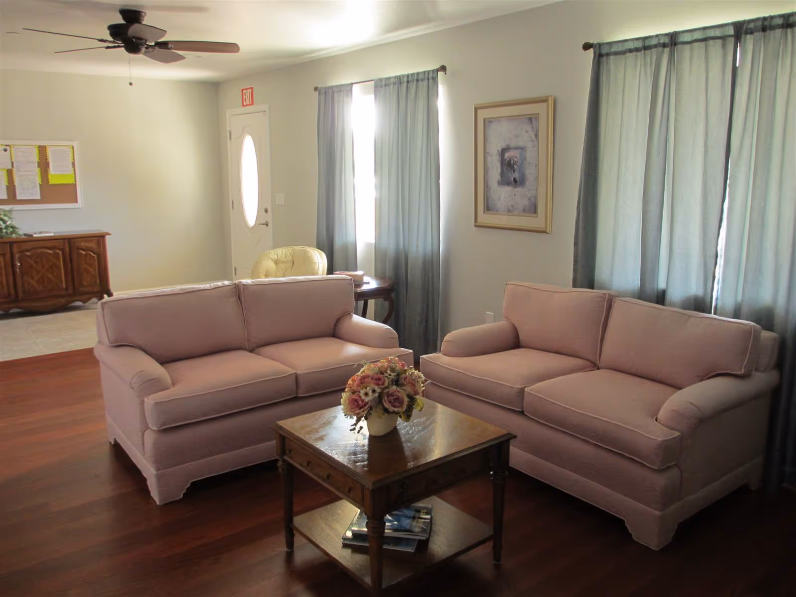 Living room with two pink sofas facing a wooden coffee table topped with a floral arrangement, hardwood floors and curtained windows.