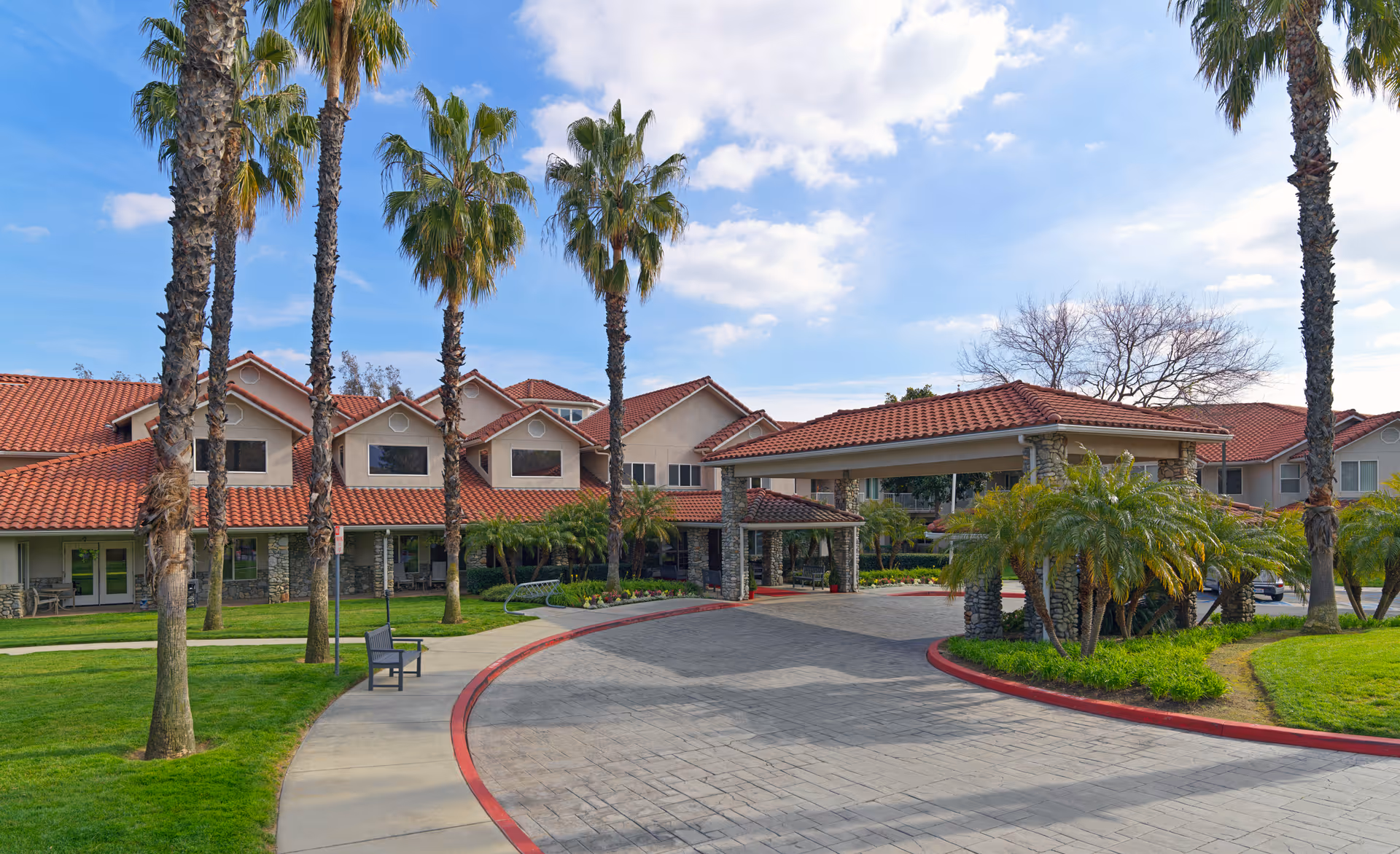 Exterior view of Holiday Valencia Commons senior living facility featuring a covered entrance with stone pillars, red-tiled roof, palm trees, green lawns, and a curved driveway under a partly cloudy sky.